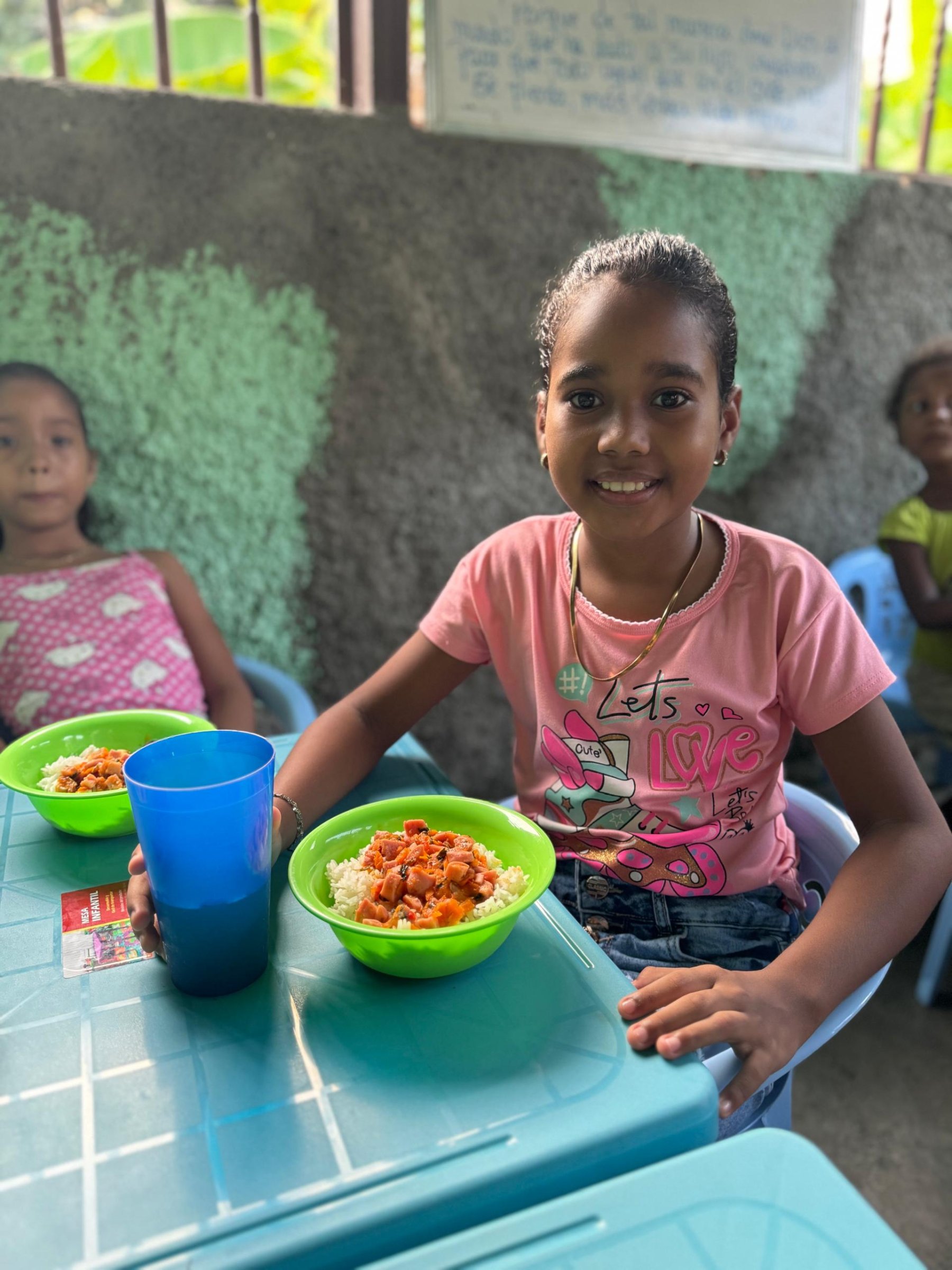 Una niña sonriente sentada a la mesa con un plato de arroz y un vaso azul.