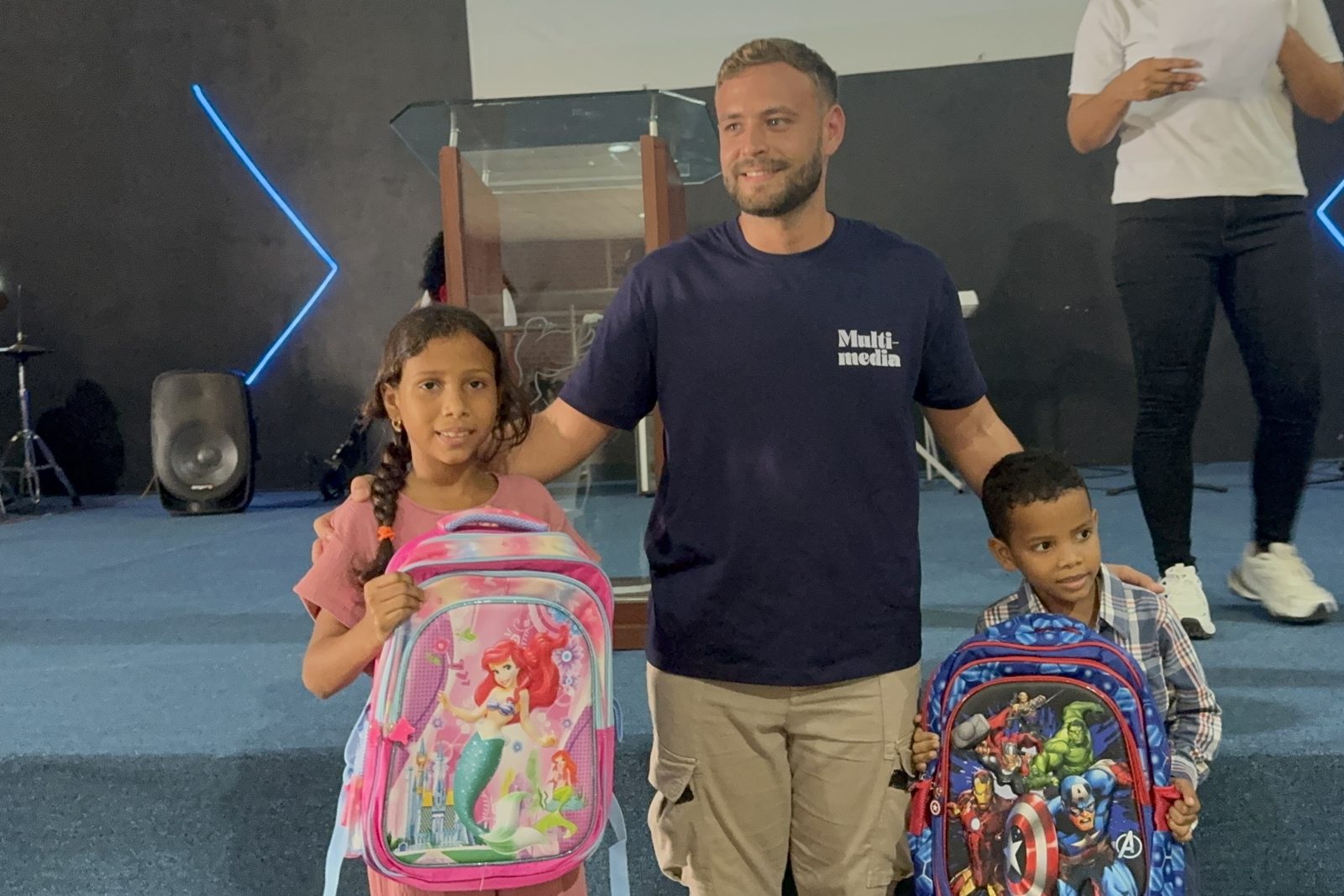 A volunteer stands with a girl and a boy holding their new school backpacks.