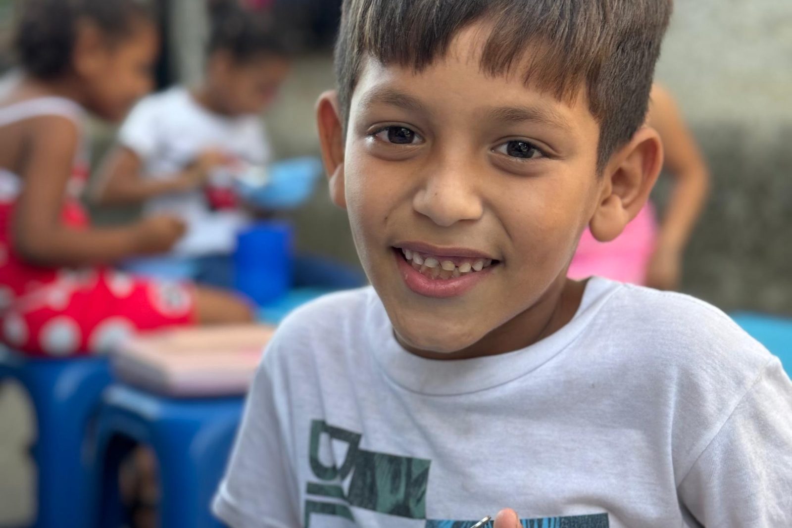 A smiling boy at a meal table, with other children eating behind him.