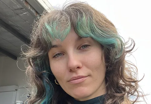 Close-up of a woman with curly brown hair and teal highlights, wearing gold hoop earrings and a black top.