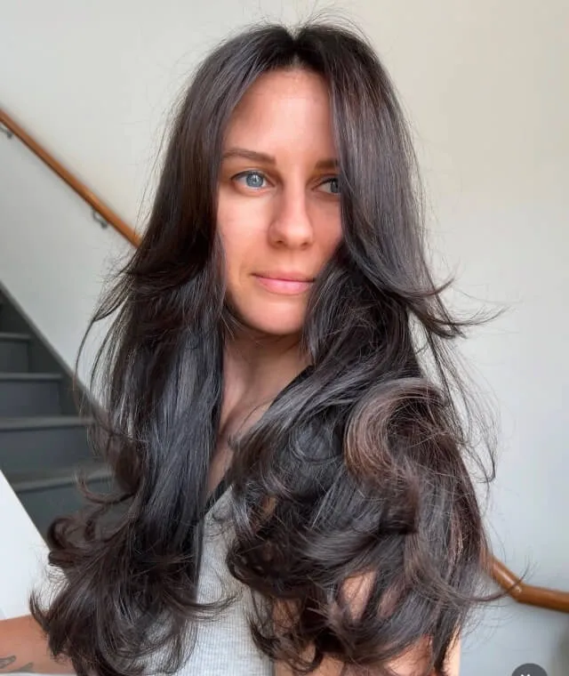 Woman with long, thick, wavy dark brown hair and blue eyes standing indoors near a staircase.