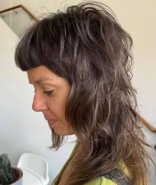Profile of a woman with thick, tousled dark hair and blunt bangs wearing a green top indoors.