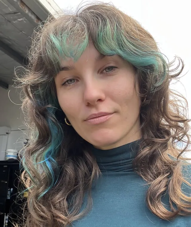 Woman with curly brown hair highlighted with teal streaks wearing a teal turtleneck, standing indoors with a neutral expression.