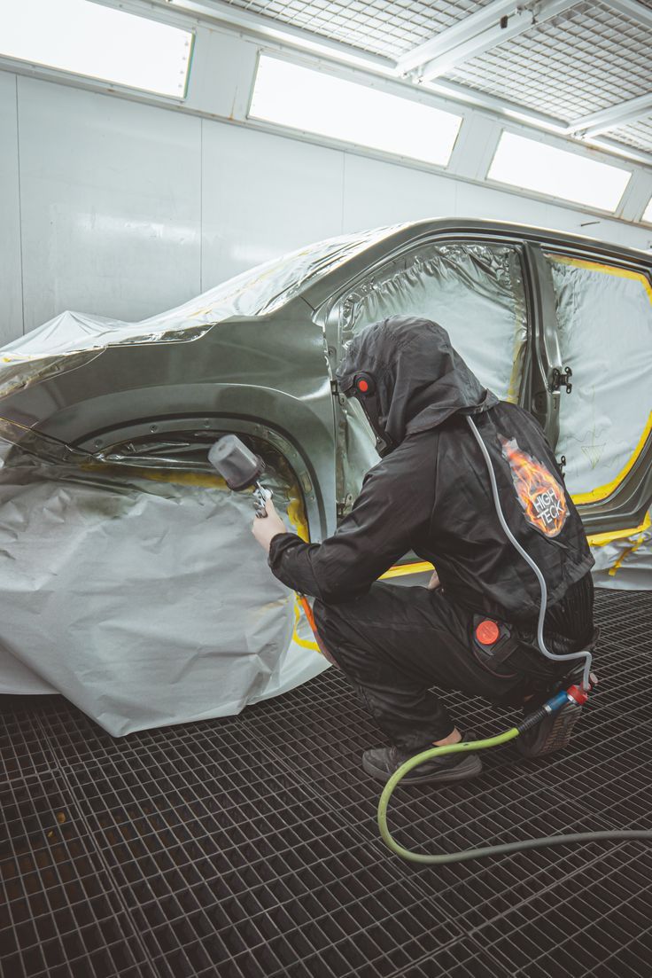 Person in protective suit painting a car fender with a spray gun in a paint booth.