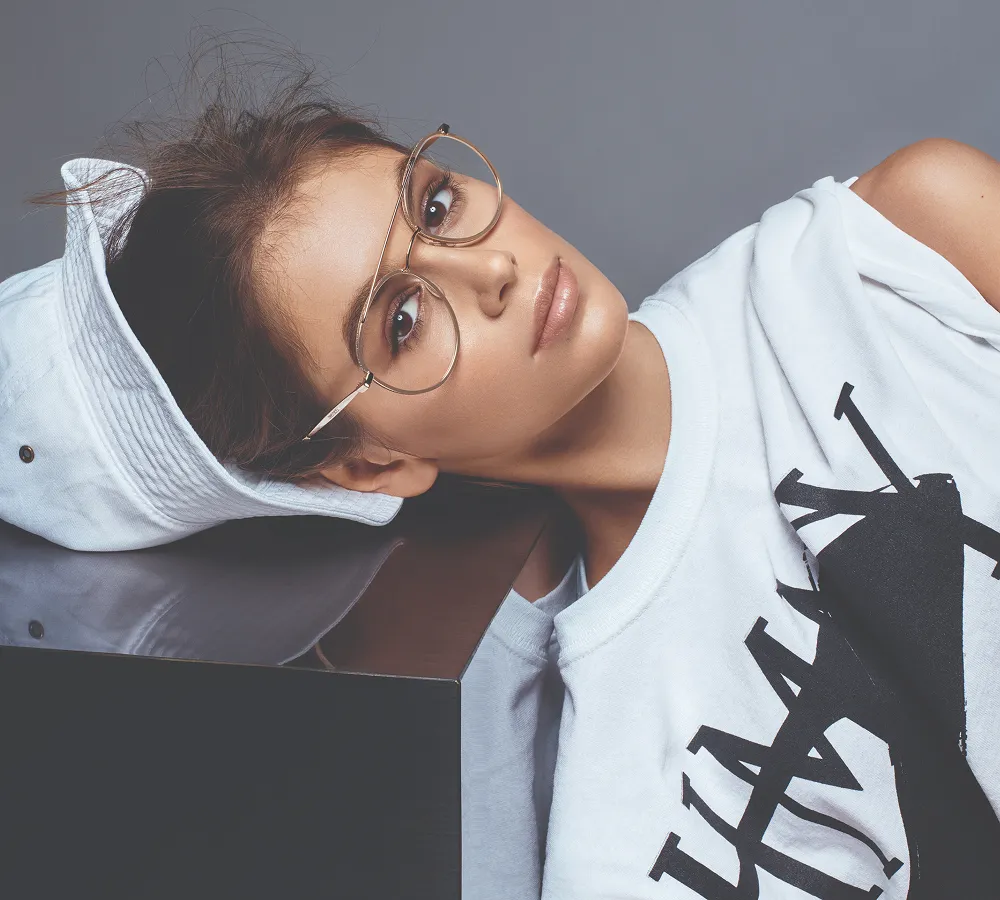Young woman wearing round glasses and a white sleeveless top, resting her head on a reflective black surface with a white hat behind her.