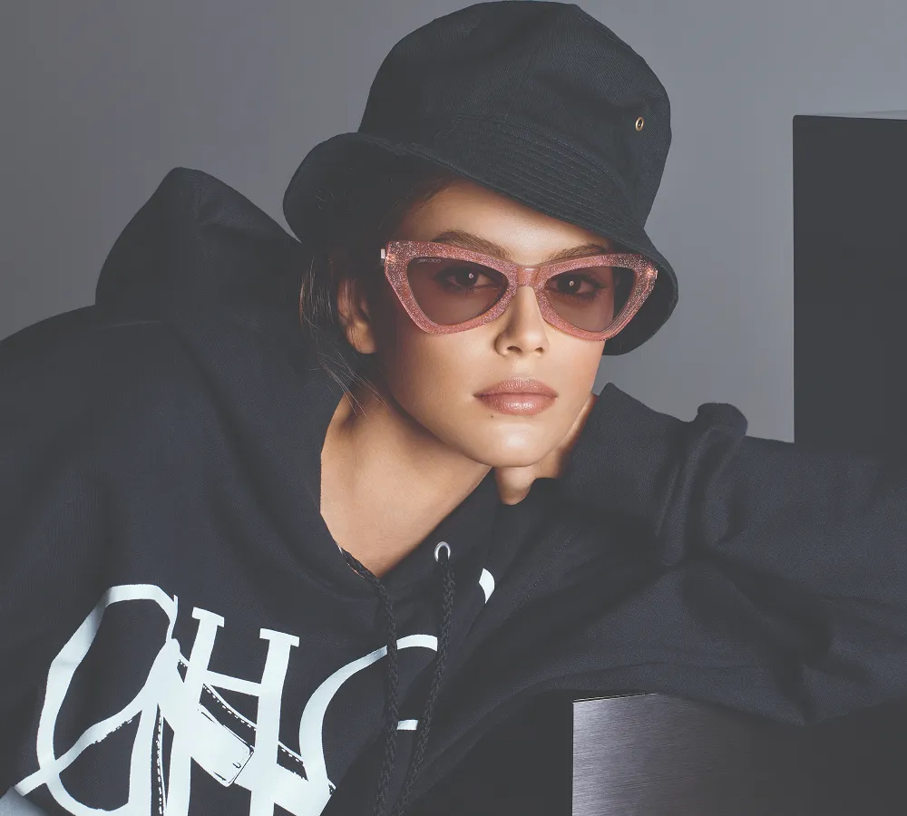 Woman wearing a black bucket hat, black hoodie with white graphic, and pink glittery cat-eye sunglasses.