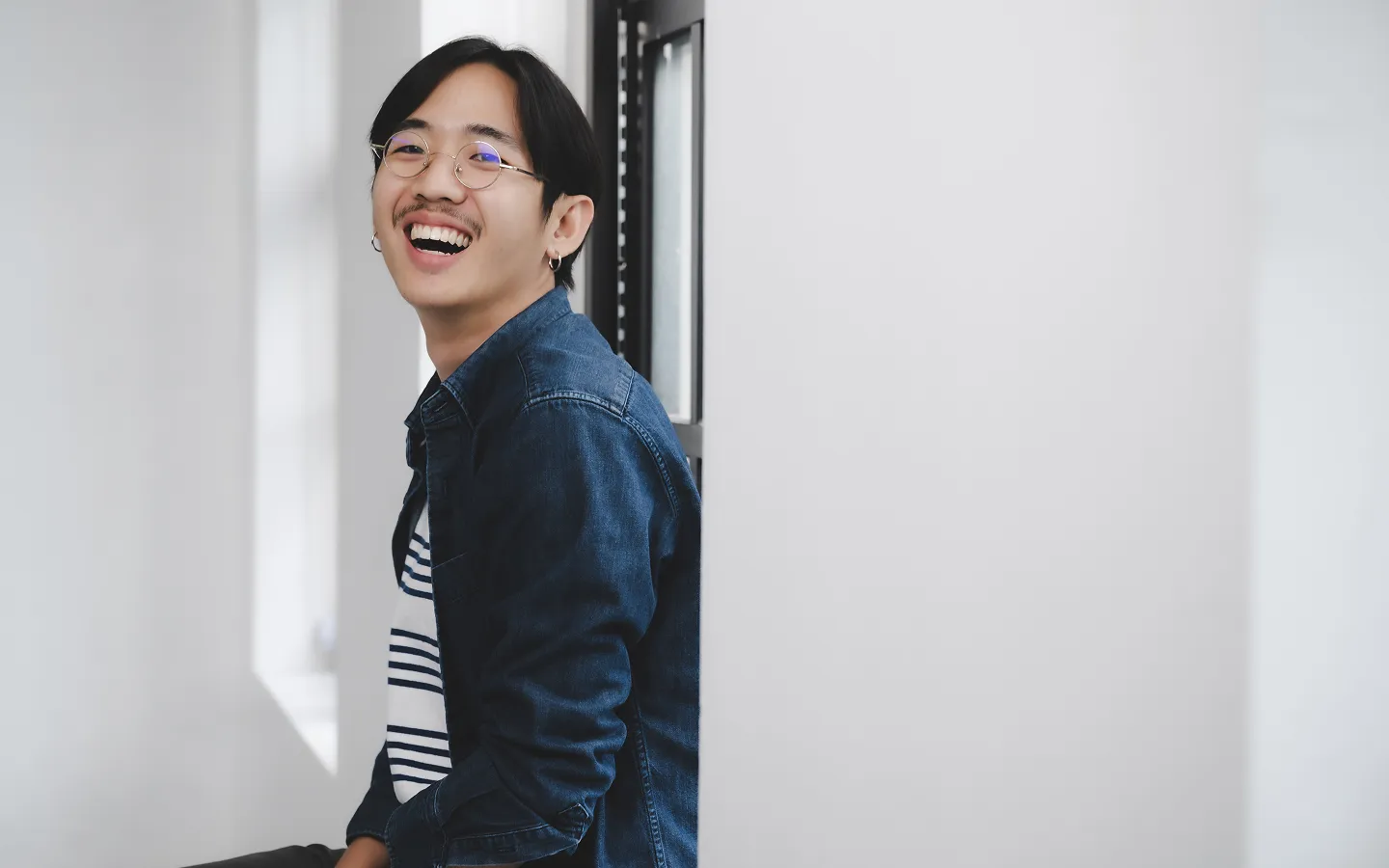 Smiling young man with glasses and earrings wearing a denim jacket and striped shirt sitting by a window.