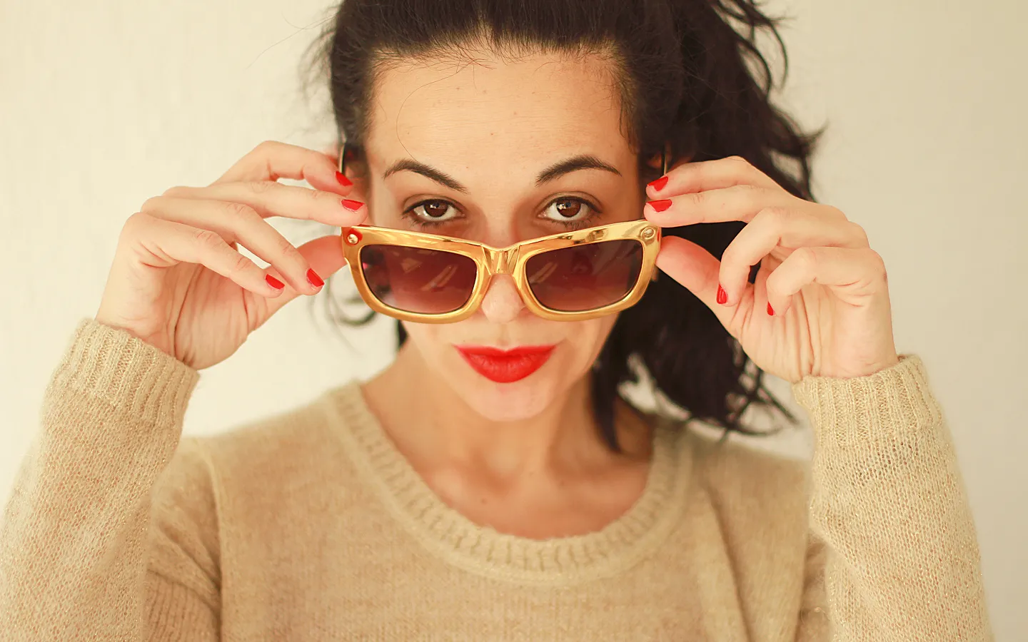 Woman with dark hair wearing red lipstick and nail polish holding golden framed sunglasses near her eyes.