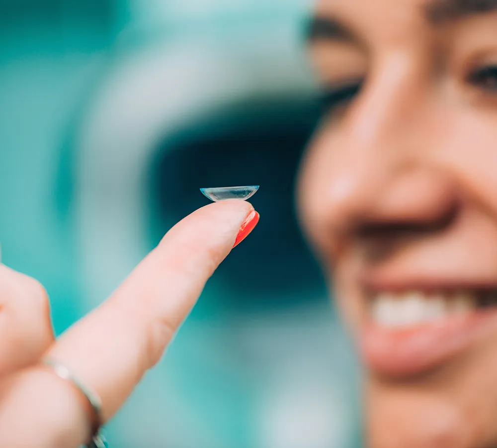 Person holding a clear contact lens on their fingertip, smiling in the background.