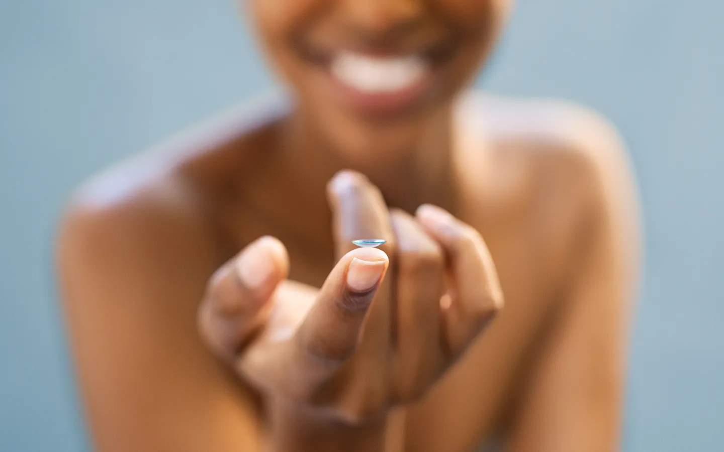 Smiling person holding a blue contact lens on their fingertip against a blurred background.