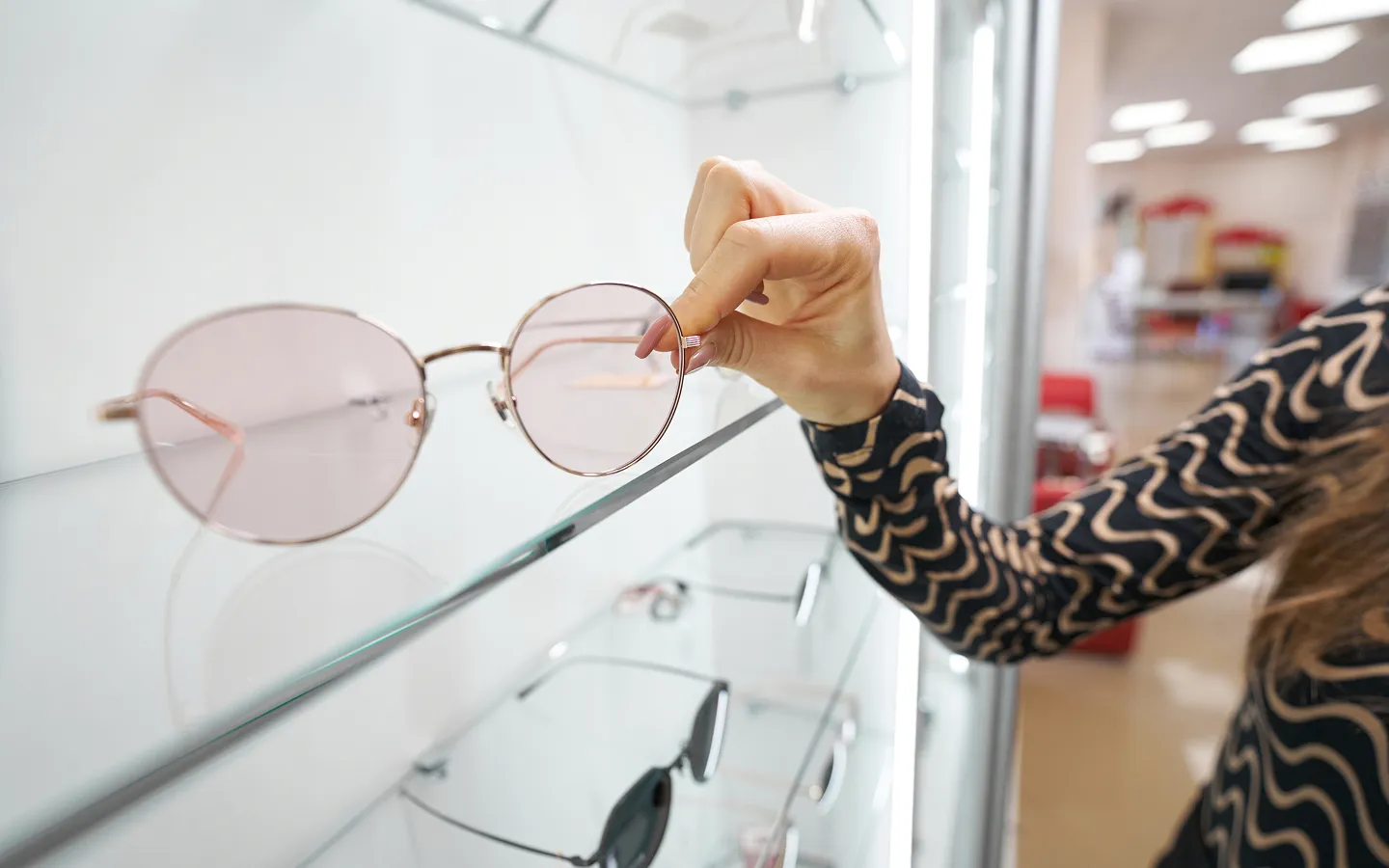 Person with patterned long-sleeve shirt holding round pink-tinted sunglasses on a glass display shelf in a store.