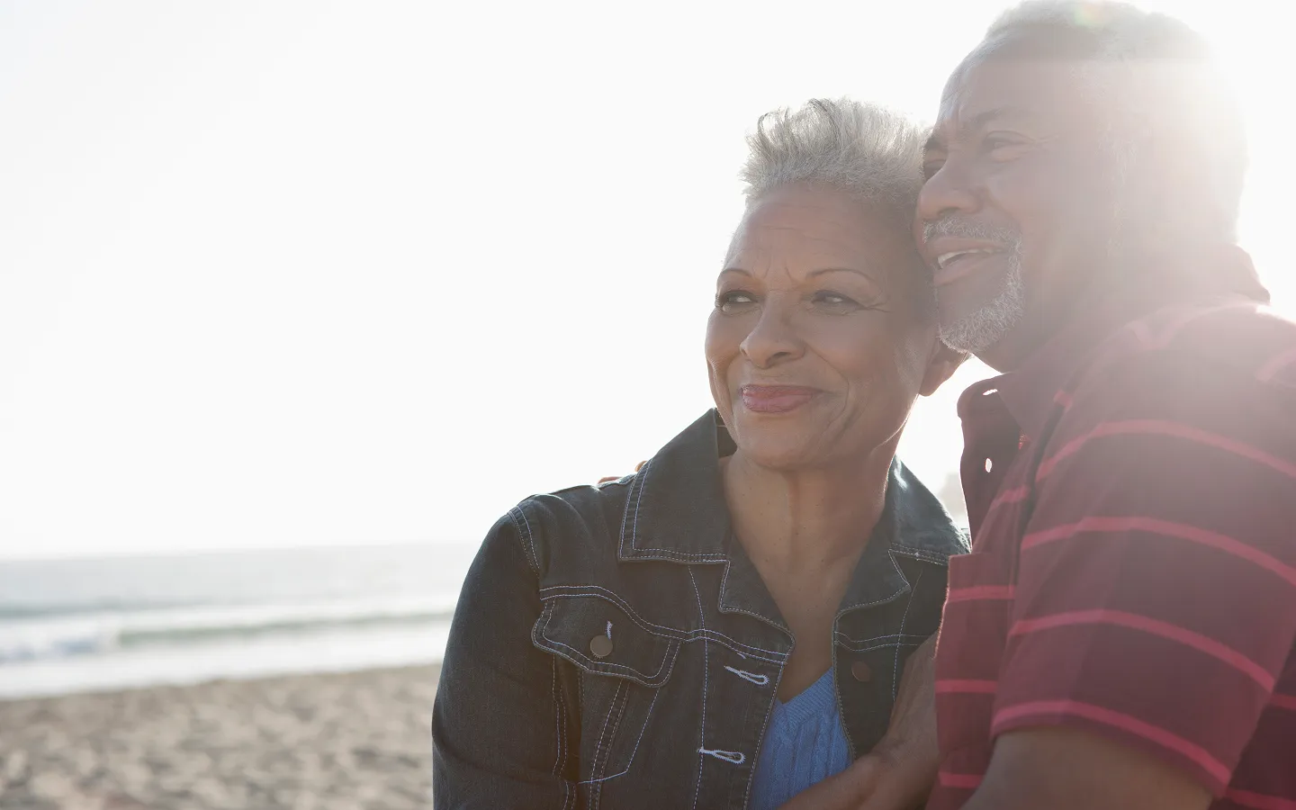 Smiling elderly couple embracing at the beach with sunlight in the background.