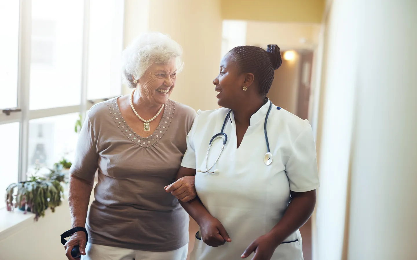 Smiling nurse with stethoscope assisting elderly woman with walking and cane in a bright hallway.