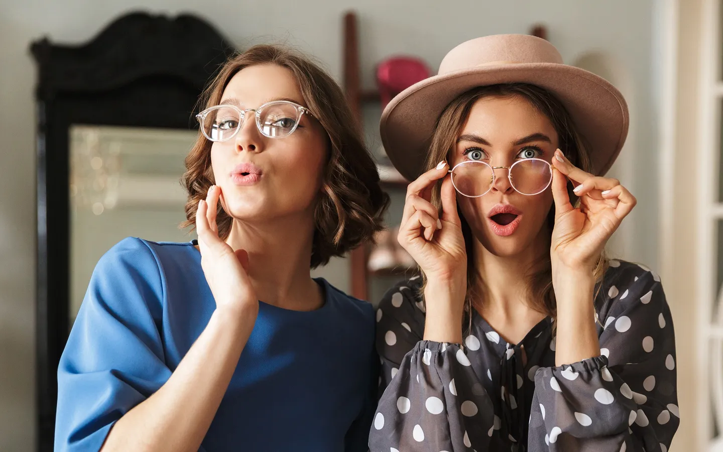Two young women wearing glasses, one in a blue top with short hair and the other in a polka-dot blouse and hat, both making surprised expressions.