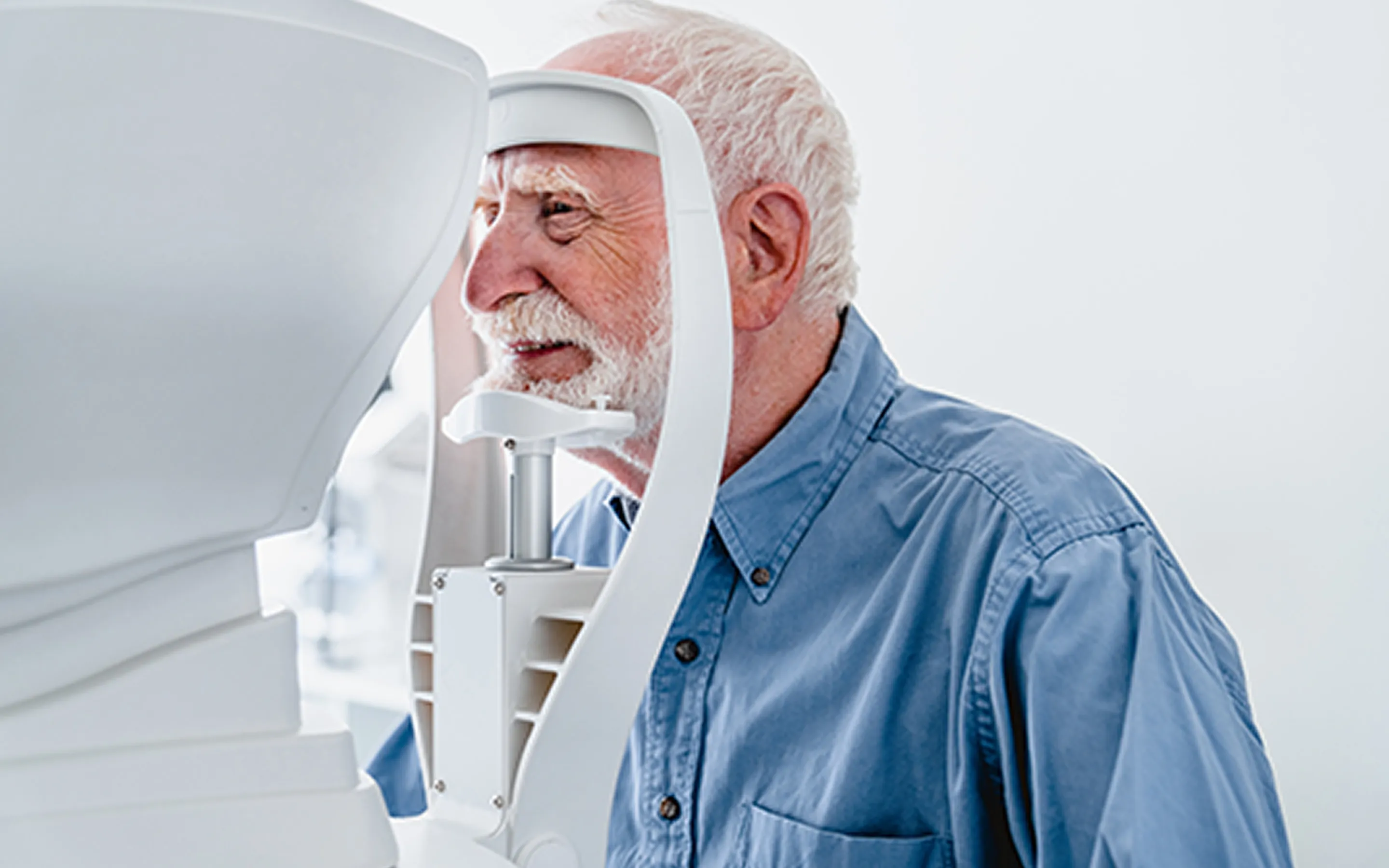 Elderly man wearing a blue shirt undergoing an eye exam using a specialized vision testing machine.