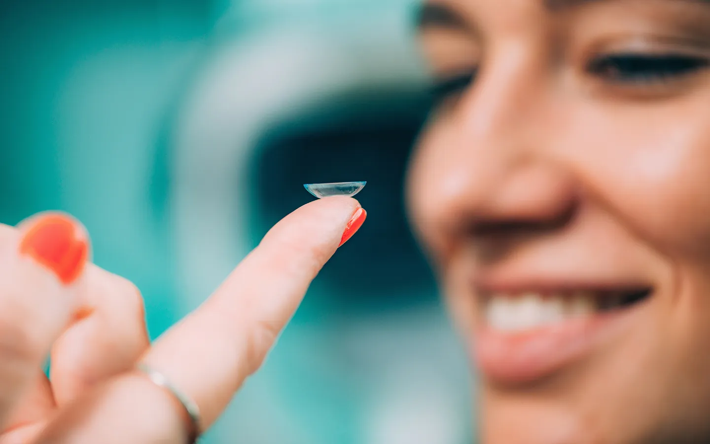A smiling woman holds a soft contact lens on the tip of her finger with red nail polish.