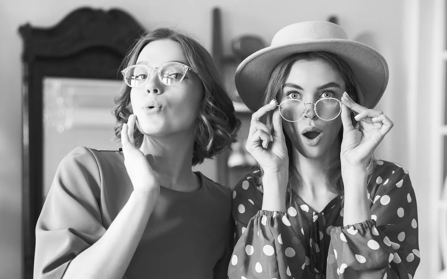 Two young women with glasses making surprised facial expressions, one wearing a hat and polka dot blouse.