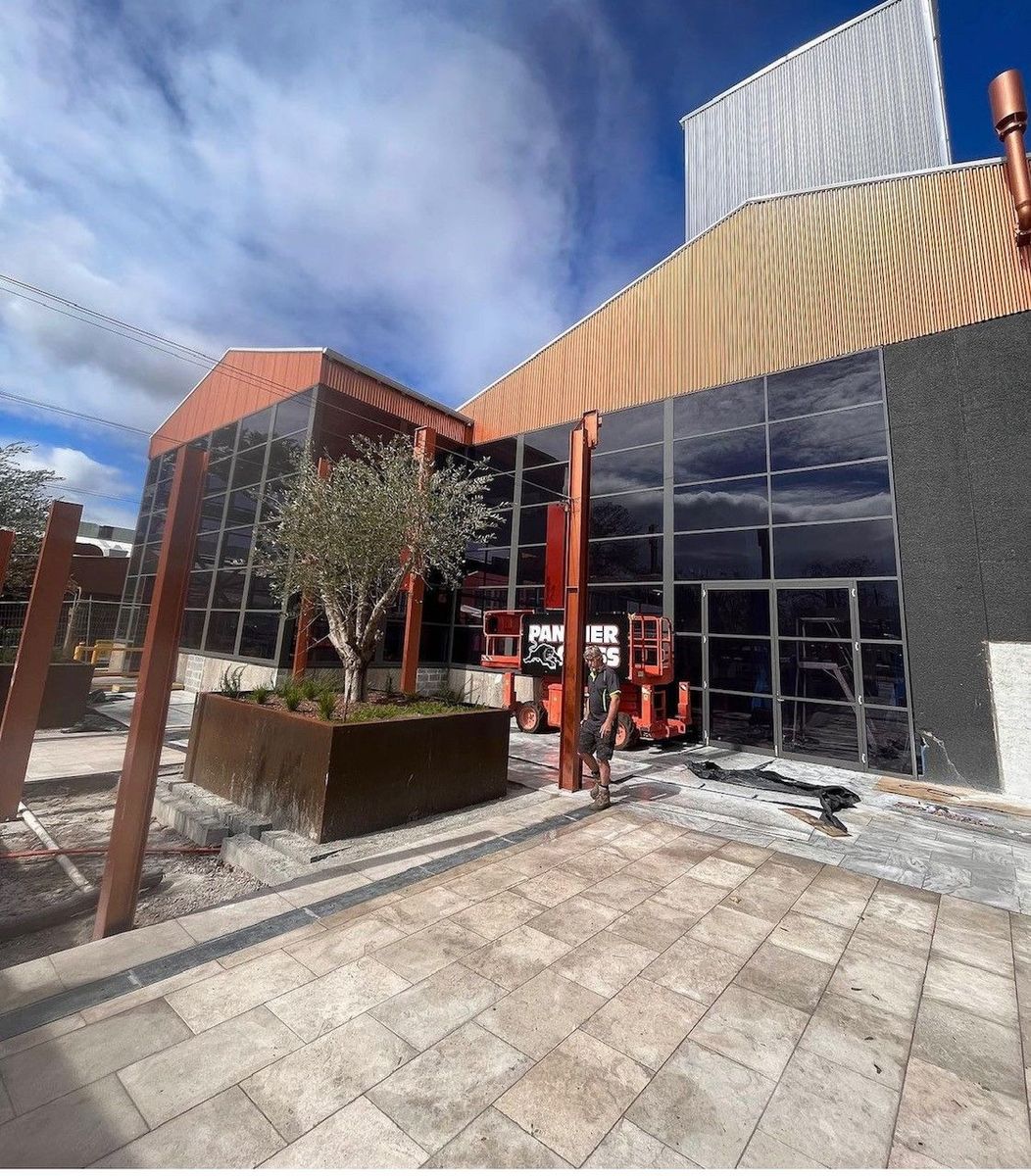 Modern industrial building with large reflective glass windows, a metal planter with a tree, and a person standing near orange construction equipment under a partly cloudy sky.