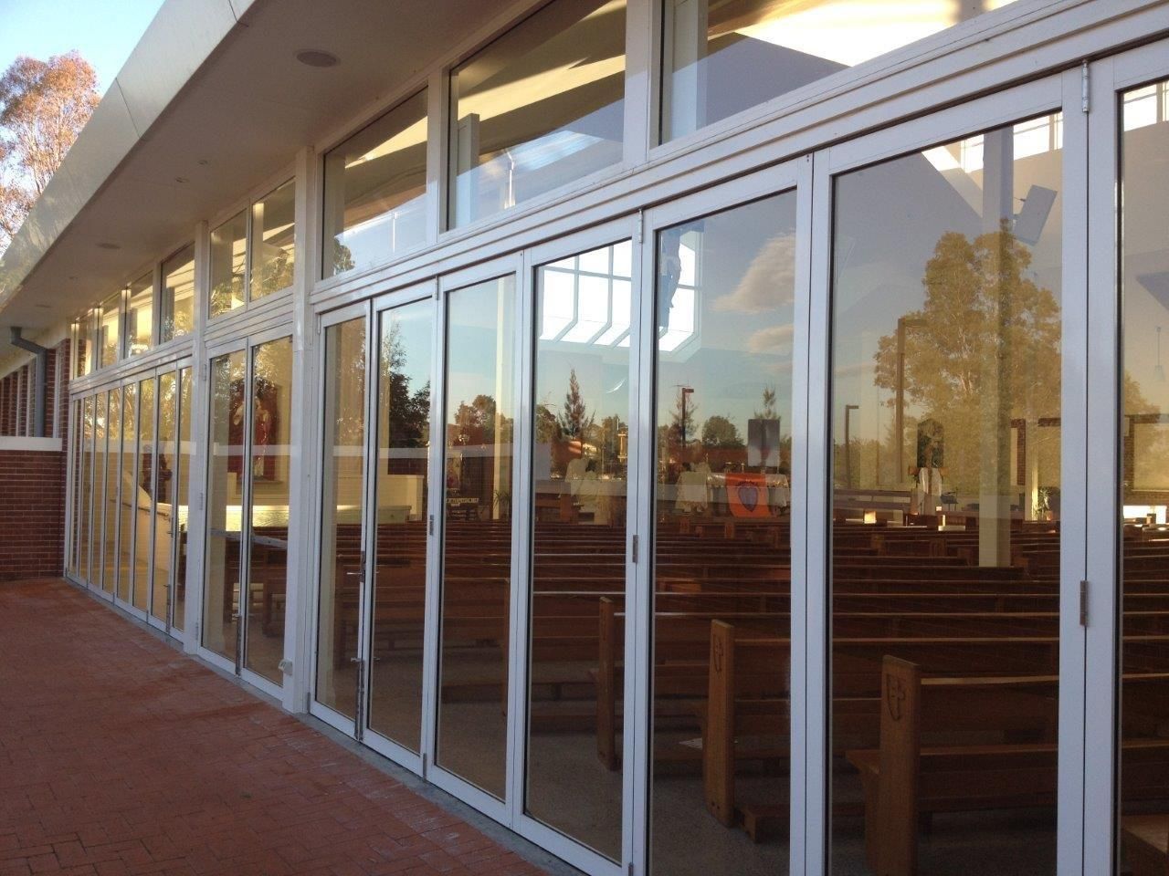 Glass exterior wall with white frames reflecting trees, showing interior wooden pews of a church.