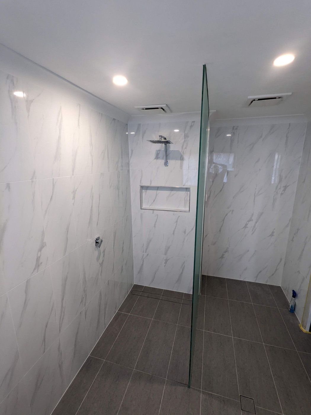 Modern frameless shower with marble-patterned wall tiles, gray floor tiles, recessed shelf, and ceiling lights.