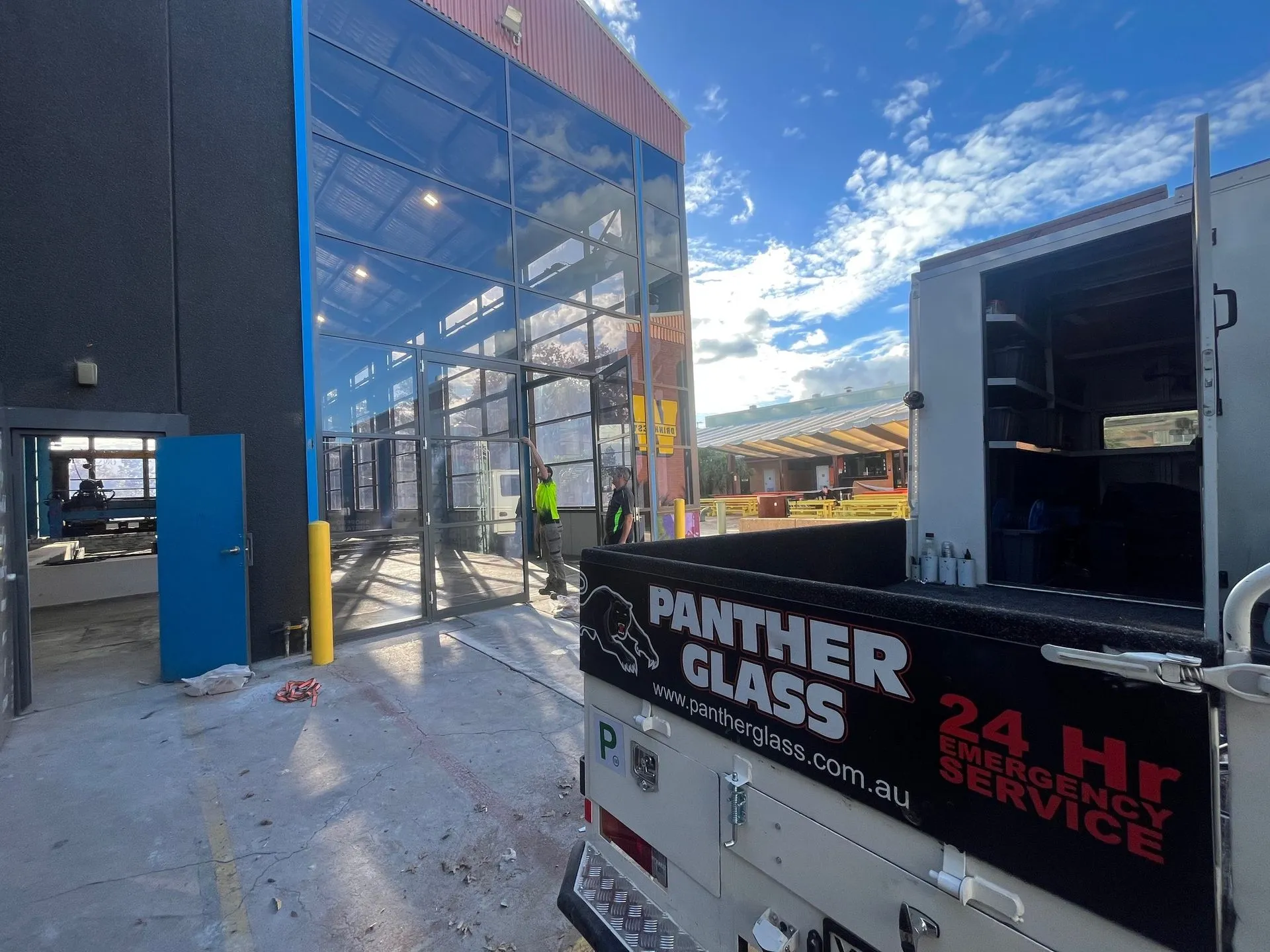 Two workers installing large glass panels on a building facade next to a Panther Glass service vehicle under a blue sky with scattered clouds.