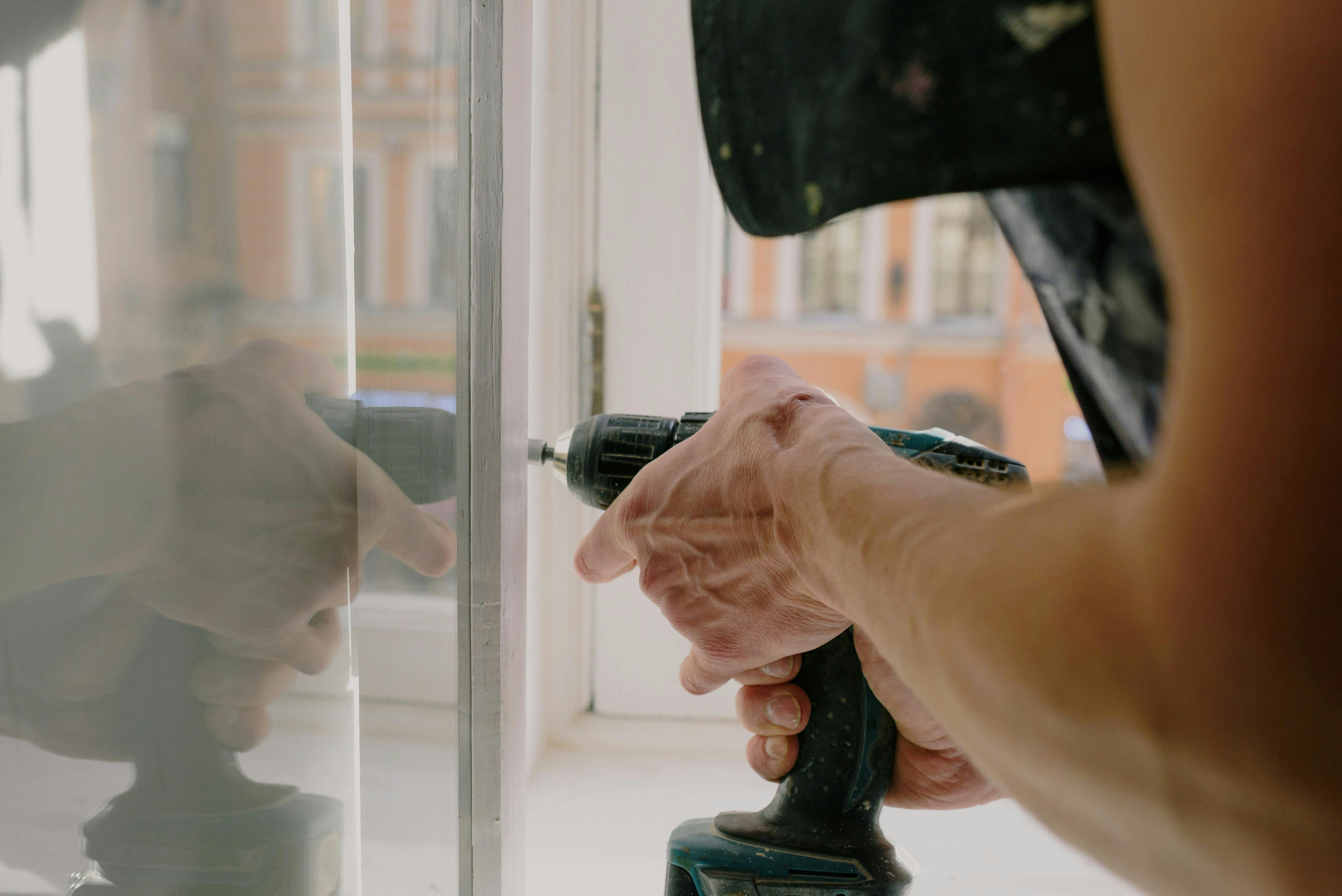 Close-up of a person using a cordless drill near a window frame with a reflection on the glass.