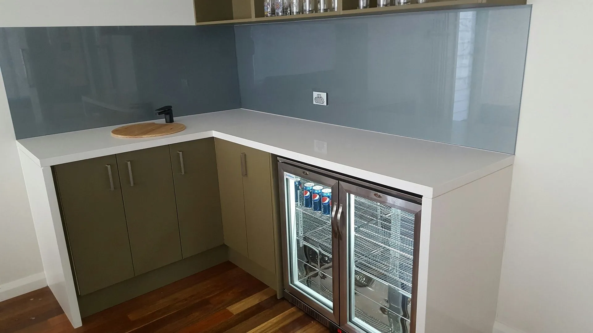 Modern corner kitchen counter with white countertop, olive green cabinets, glass backsplash, black faucet, wooden cutting board, and a double-door glass refrigerator containing Pepsi cans.