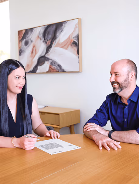 A woman with long black hair holding a pen signs a document on a wooden table while a bald man with a beard in a blue shirt smiles at her.