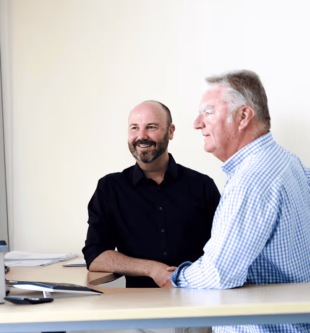 Two men sitting at a desk, one in a black shirt smiling and facing forward, the other in a blue checkered shirt looking to the side.