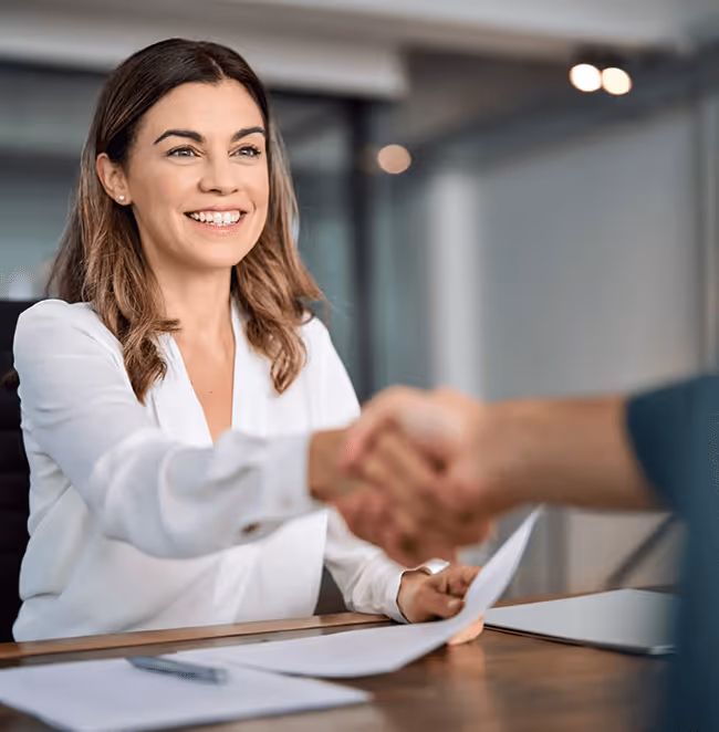 Smiling woman in white blouse shaking hands with another person across a desk in an office setting.