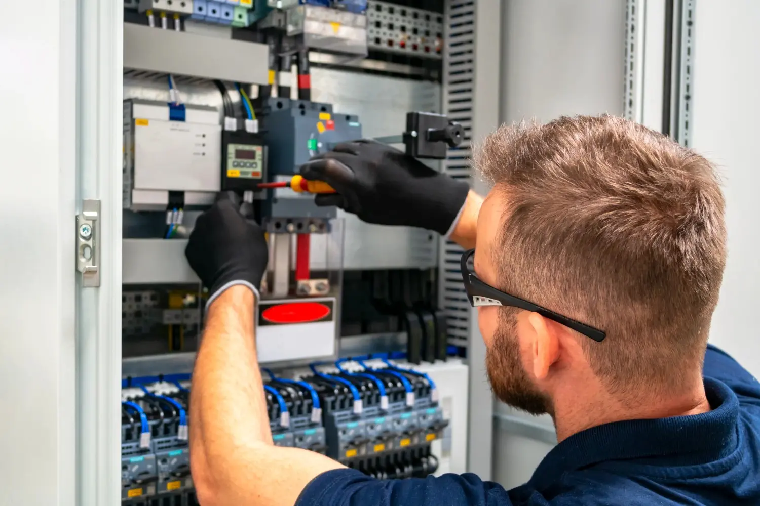 A technician wearing black safety gloves performs maintenance on a complex industrial control panel with wiring