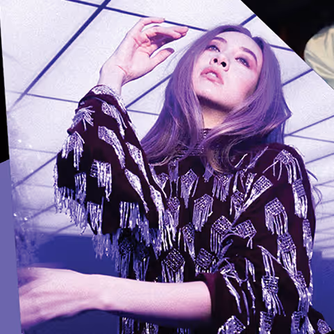 Young woman with long purple hair wearing a black and silver fringed outfit posing dramatically under a tiled ceiling.