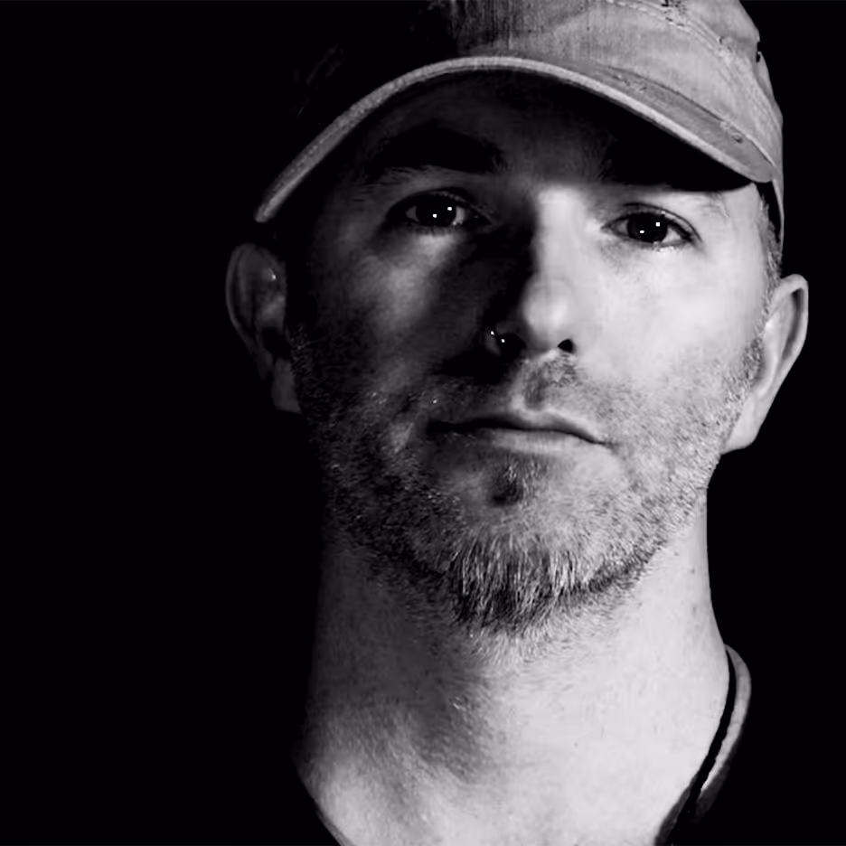 Black and white portrait of a man with a trimmed beard wearing a baseball cap against a dark background.