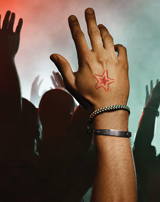 Raised hand with a red star tattoo and bracelets against a crowd with raised hands at a concert with red and green lighting.