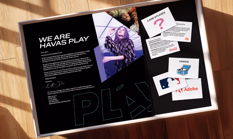 A framed black presentation board on a wooden floor with the title 'We Are Havas Play,' a letter from the VP, an image of a woman posing, and cards showing case studies and credits including Coca-Cola, MLB, Adobe, and JBL logos.