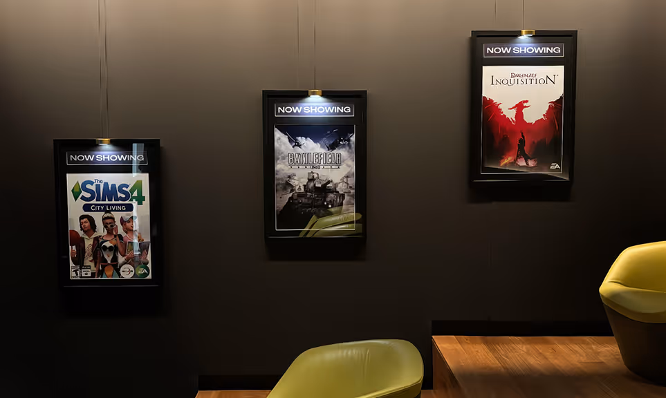 Three framed video game posters labeled 'Now Showing' on a dark wall above green chairs and a wooden table.