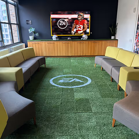 Modern office lounge with green carpet featuring EA logo, gray and green couches, and a TV displaying an EA Sports football player.