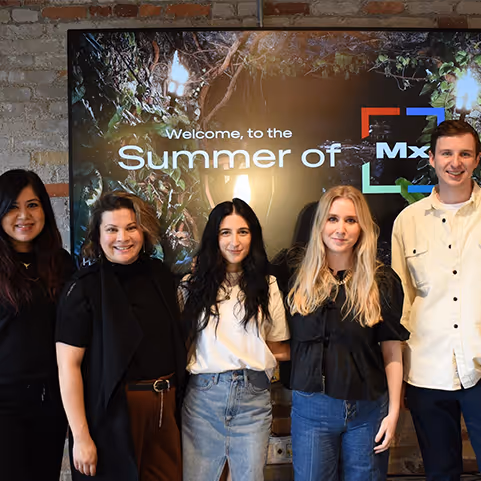 Five diverse people standing shoulder to shoulder smiling in front of a screen with the text 'Welcome to the Summer of Mx'.