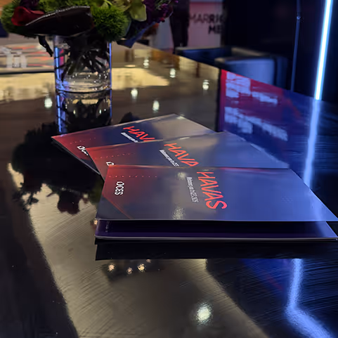 Stack of magazines titled 'HAVAS' on a reflective dark table with a glass vase of flowers in the background.