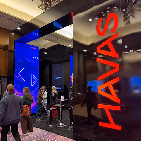 People walking and standing near a booth with large digital displays, one showing the word 'HAVAS' in red letters.