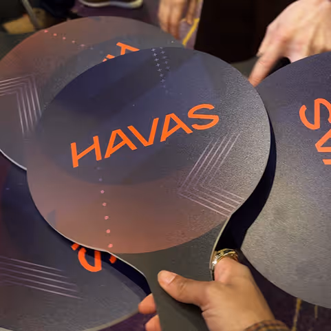 Hand holding a black ping pong paddle with the word 'HAVAS' printed in orange on it.