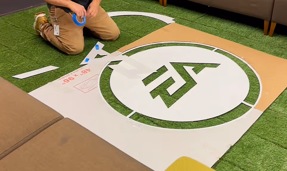 Person kneeling on artificial grass taping paper stencil with the EA logo cut out on the floor.