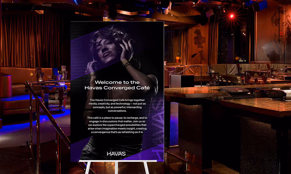 Dimly lit lounge area with tables and chairs, featuring a large vertical poster with a woman listening to music and text about the Havas Converged Café.
