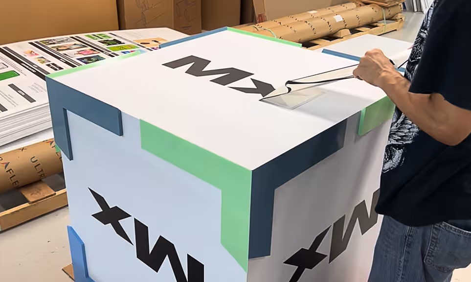 Person laminating a cardboard box with black text 'Mx' printed on its sides, in a workshop setting.