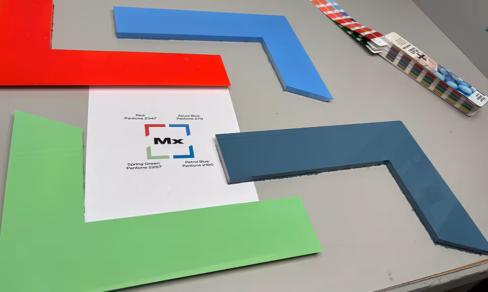 Four large L-shaped color samples in red, blue, green, and dark blue surround a paper with color names and Pantone codes on a gray surface with a Pantone color guide fan nearby.