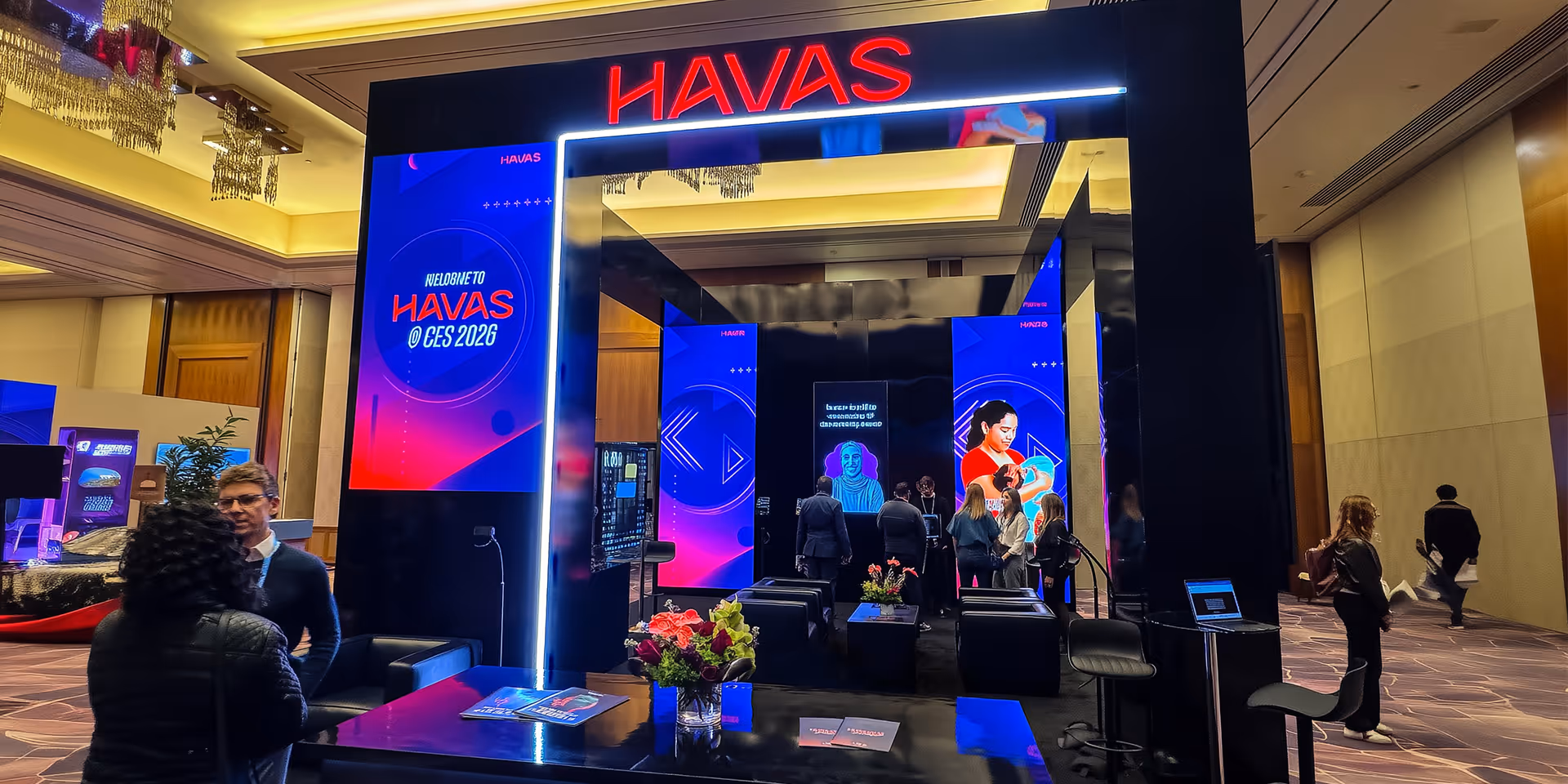 Exhibition booth with large illuminated HAVAS sign and digital screens displaying CES 2026 branding with people interacting inside.