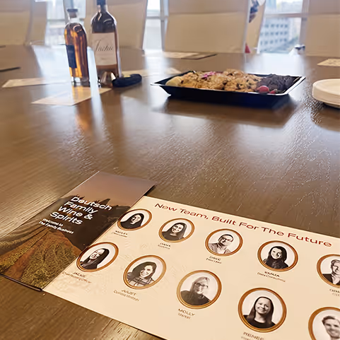 Brochure displaying the new team of Deutsch Family Wine & Spirits on a wooden conference table with bottles and a tray of cookies in the background.