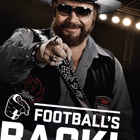 Man wearing sunglasses, a hat, and a vest pointing forward with a confident smile against a dark background with stage lights.