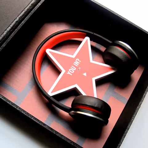 Black and red over-ear headphones resting inside a black box with a red star-shaped card that reads 'YOU IN?'.