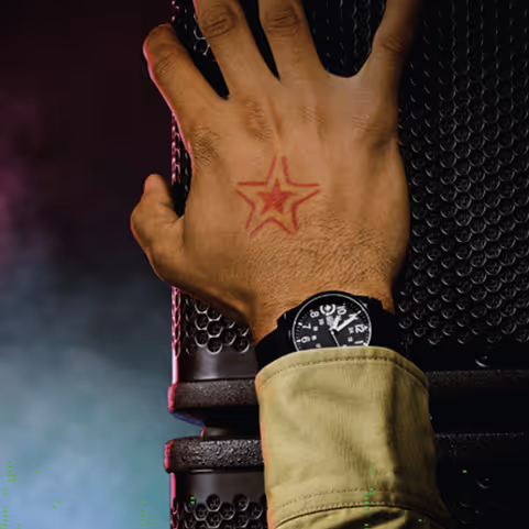 Close-up of a hand with a red star tattoo placed on a black textured surface, wrist wearing a black watch and a greenish sleeve.